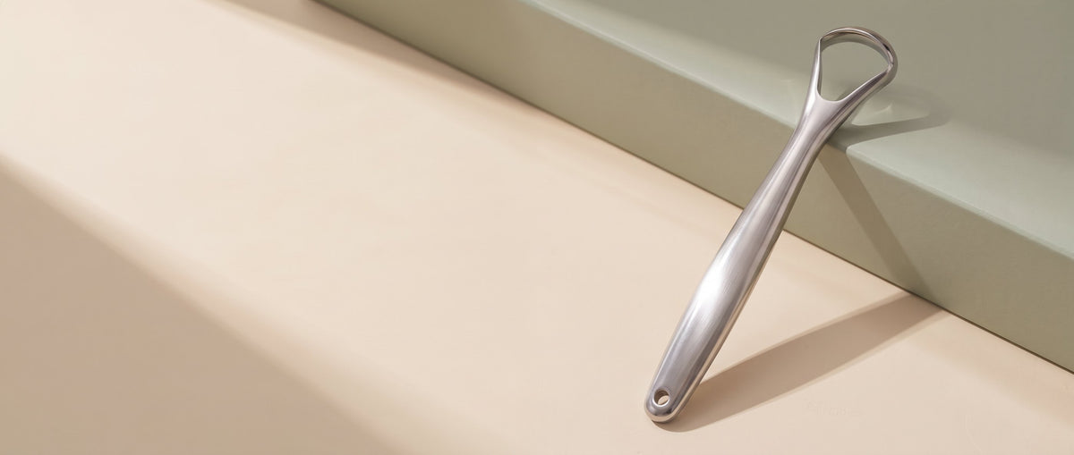 Stainless-steel OralCove tongue scraper standing on the right side of a warm neutral scene.